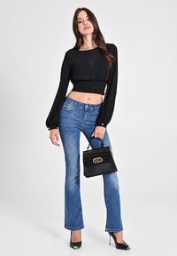 Schwarzes langärmliges, bauchfreies Oberteil, blaue weit fallende Jeans, hält eine schwarze Handtasche im Krokodil-Muster mit goldenen Beschlägen, kombiniert mit schwarzen Absätzen.