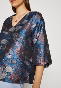 Blumenmuster-Bluse mit V-Ausschnitt und kurzen Puffärmeln. Aus metallisch schimmerndem lila und blauen Stoff mit strukturierter Oberfläche und grauen Akzenten.