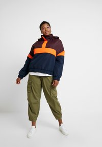Chaqueta a bloques de color con paneles en burdeos, naranja y marino; puños elásticos; diseño con capucha; combinada con pantalones cargo oliva y zapatillas blancas.