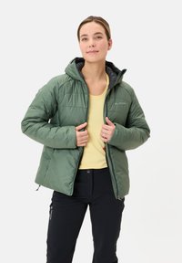 Giacca isolante verde con cappuccio, motivo trapuntato e chiusura con zip. Indossata sopra una maglietta gialla, con pantaloni neri visibili.