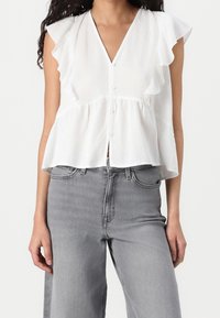 Wit, kortgeleden top met ruches, knoopsluiting en een casual pasvorm. Gecombineerd met lichtgrijze wijde jeans.