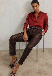 Punainen satiininen nappipaita ja ruskeat metallic-korkeavyötäröiset housut, yhdistettynä mustiin korkokenkiin. Istuu kirkkaalla tuolilla.