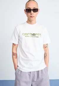 Witte t-shirt met korte mouwen met groene "Carhartt Work in Progress" opdruk. Draag het met een lichtgrijze short met patroon. Soepele stof, relaxte pasvorm.