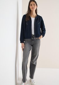 Veste à capuche bleu marine avec poignets élastiques, fermeture éclair et poches avant, associée à un jean gris droit et des baskets blanches.