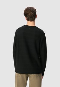 Uomo con capelli corti castani che indossa una maglietta nera a maniche lunghe con texture e pantaloni beige, di spalle contro uno sfondo chiaro e semplice.