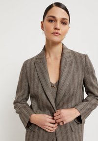Bruine pinstripe blazer met een notch revers, enkele knoopsluiting en gestructureerde stof. Gepaard met een gelaagd top en ringen.