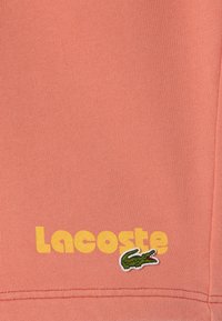 Sudadera de algodón color coral con un logo amarillo de "Lacoste" y un acento de cocodrilo bordado en verde. Textura suave con detalles acanalados.