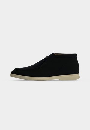 Chaussure décontractée mid-top noire avec semelle beige, bout style mocassin et détail subtil de laçage sur un fond clair uni.