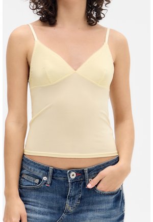 Woman wearing a pale yellow fitted camisole with thin straps and blue denim jeans, one hand partially in pocket.