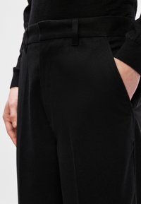 Calças pretas ajustadas com uma textura suave, apresentando bolsos laterais e uma cintura plana na frente. Detalhe de costura visível na cintura.