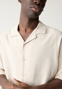 Camicia beige a maniche corte in lino leggero. Presenta un colletto alla cubana, una texture sottile e una chiusura frontale con due bottoni.