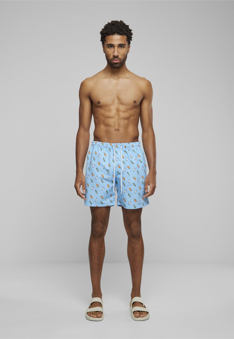 Homme aux cheveux bouclés portant un short de bain bleu à motif de sucettes glacées et des sandales blanches, debout devant un fond gris clair.