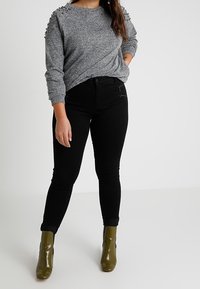 Grijze gebreide trui met versierde schouders, gecombineerd met zwarte skinny jeans en olijfgroene enkellaarzen. Eenvoudig, modern ontwerp.