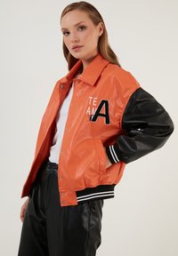 Oranje faux leren varsityjack met zwarte leren mouwen, geribbelde manchetten en borduursel "TEAM A" op de voorkant.