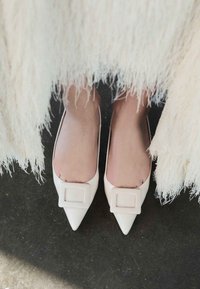 Picioare purtând pantofi albi cu vârf ascuțit și catarame pătrate, parțial acoperiți de un articol vestimentar lung și texturat, stând pe un podea întunecată.