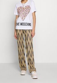 Osoba v bílém tričku s růžovým leopardím srdcem a nápisem "LOVE MOSCHINO", vzorovaných širokých kalhotách ve tvaru cik-cak, bílých teniskách a zlatém náhrdelníku.