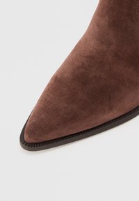 Botín de ante marrón hasta el tobillo con punta afilada, superficie texturizada y suela de goma negra contrastante. Parte superior suave, con un diseño simple y funcional.