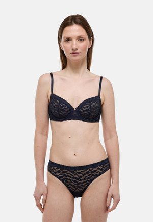 Femme debout, de face, portant un soutien-gorge en dentelle noire et une culotte assortie, sur fond uni gris clair.