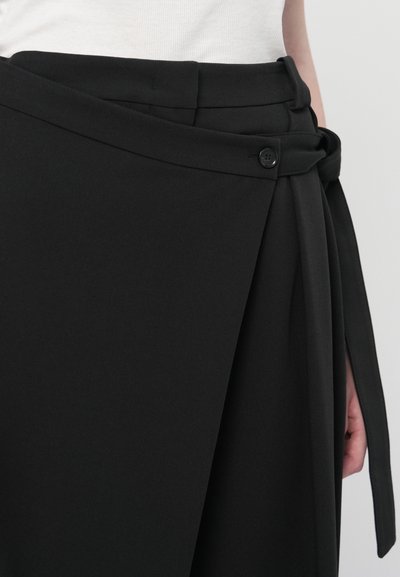 Gros plan sur une jupe noire enveloppante avec des détails de boutons et une ceinture, portée avec un haut blanc sur un fond clair.
