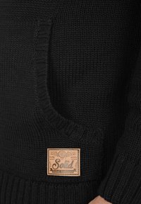 Suéter negro de punto con un bolsillo delantero, dobladillo y puños acanalados, y una etiqueta de cuero marrón claro con el nombre de la marca "Solid".