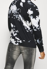 Sudadera de teñido anudado en blanco y negro con mangas largas y dobladillo acanalado, combinada con jeans grises que presentan sutiles adornos en los bolsillos.