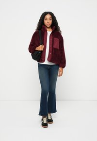 Burgunder fleecejakke med knaplukning og brystlomme, sammen med hvid skjorte, flagrende blå jeans og sorte sneakers. Sort taske over skulderen.