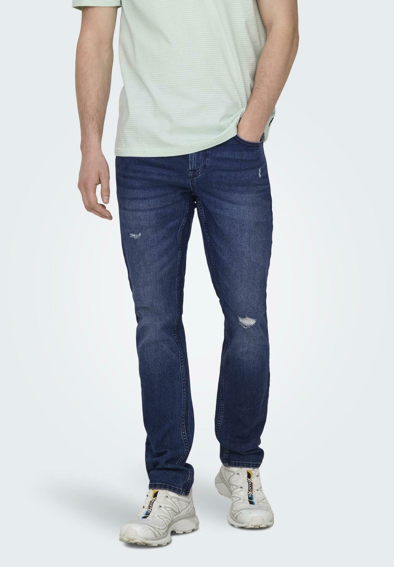 Mørkeblå jeans med falmede slidte områder og diskret whiskering. Har en lige pasform og er parret med en lysegrøn stribet skjorte.