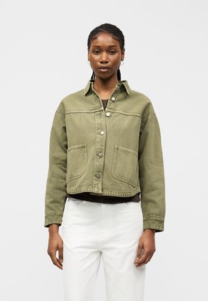 Femme aux cheveux tressés portant une veste boutonnée vert olive et un pantalon blanc, debout devant un fond clair uni.