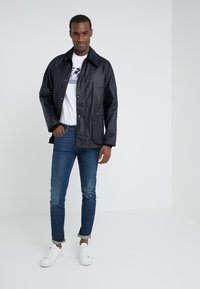 Zwarte waterdichte jas met een marineblauwe kraag, knoopsluiting aan de voorkant en borstzakken, gecombineerd met aansluitende blauwe jeans en witte sneakers.