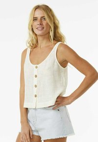 Top corto de lino blanco con tirantes anchos y escote en V, que cuenta con cinco botones de madera en el frontal. Combinado con pantalones cortos de mezclilla claros.