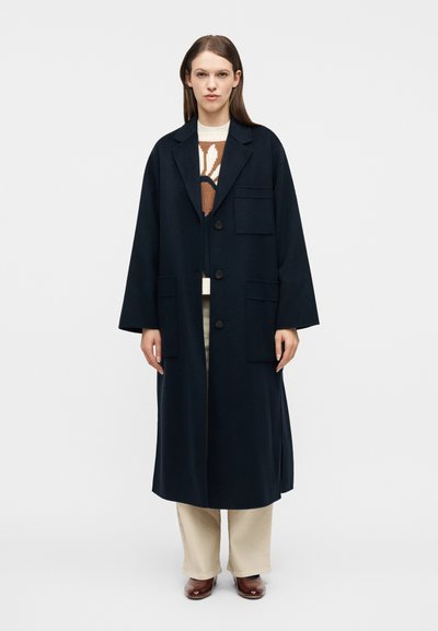Femme debout portant un long manteau bleu marine à trois boutons par-dessus un pull marron et blanc, un pantalon beige et des chaussures marron.