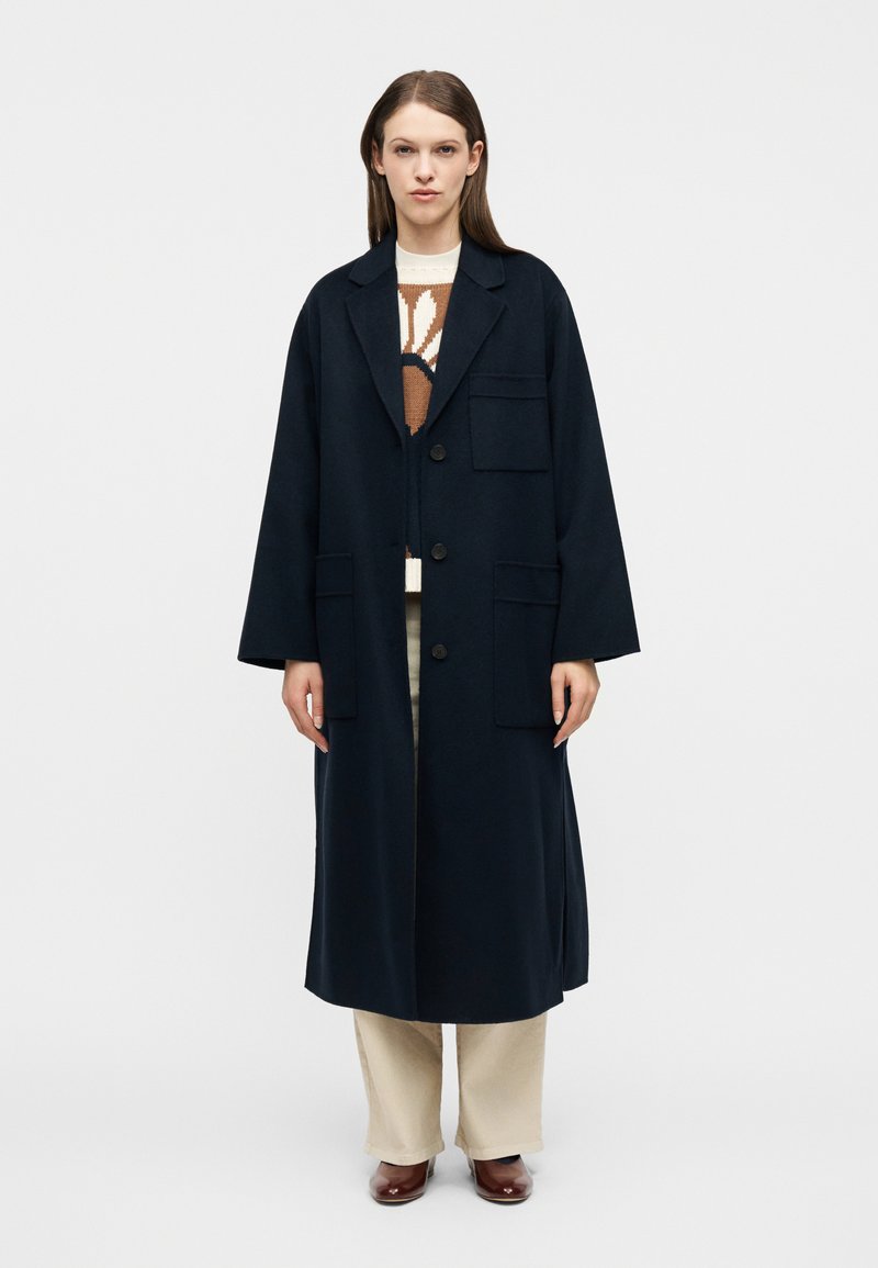 Γυναίκα που στέκεται φορώντας ένα  μακρύ μπλε παλτό με τρία κουμπιά πάνω από ένα καφέ και λευκό πουλόβερ, μπεζ παντελόνια και καφέ παπούτσια.