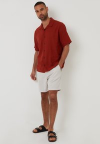 Rød linned kortærmet skjorte med sorte knapper, parret med lysegrå shorts og sorte sandaler med dobbelte stropper. Ensfarvet baggrund.