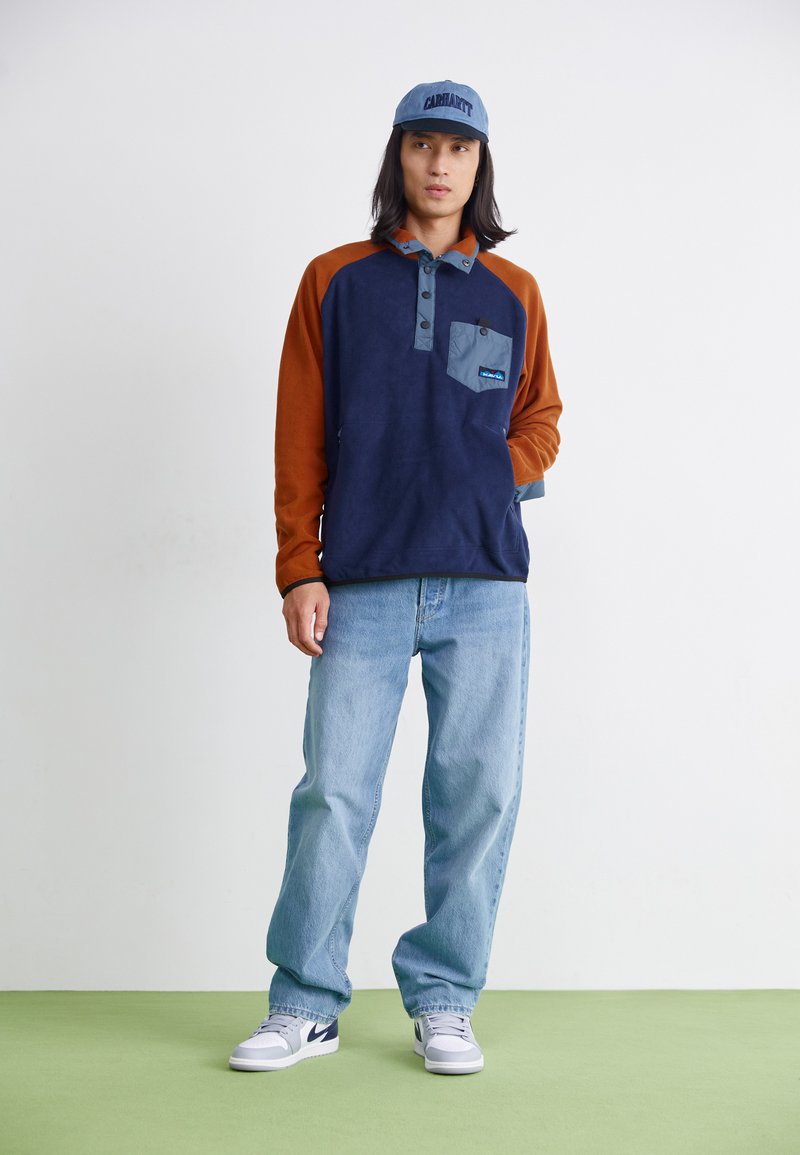 Sudadera de polar en navy y naranja con un bolsillo en el pecho, cuello abotonado gris, combinada con jeans azul claro y zapatillas blancas.