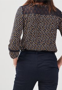 Navy blouse with a geometric pattern in orange and cream; features sheer lace back yoke, roll-up sleeves, and a relaxed fit.