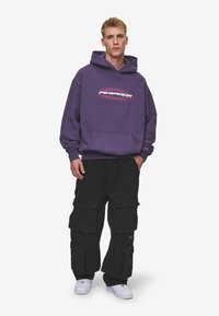 Hanorac mov cu logo frontal, buzunar tip kangaroo, asortat cu pantaloni cargo negri cu multiple buzunare. Adidași albi completează look-ul.