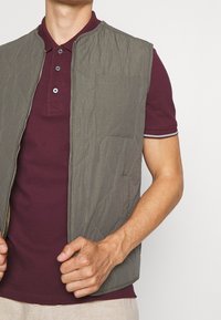 Grå vadderad väst med dragkedja framtill, med en bröstficka, som bärs över en vinröd polo t-shirt med knäppning med tre knappar och kontrasterande kant.