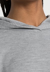 Nahaufnahme eines Nackens und der Schultern einer Person, die einen hellgrauen, gerippten Kapuzenpullover trägt, mit geflochtenem Haar, das seitlich sichtbar ist.