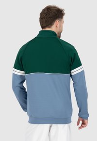 Giacca a pullover con colletto alto, caratterizzata da una parte superiore verde, una parte inferiore blu chiaro e strisce bianche sulle maniche. Texture del tessuto morbida.