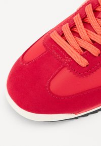 Red athletic shoe featuring a mix of suede and synthetic materials, with orange laces and accents, rounded toe, and a white sole.