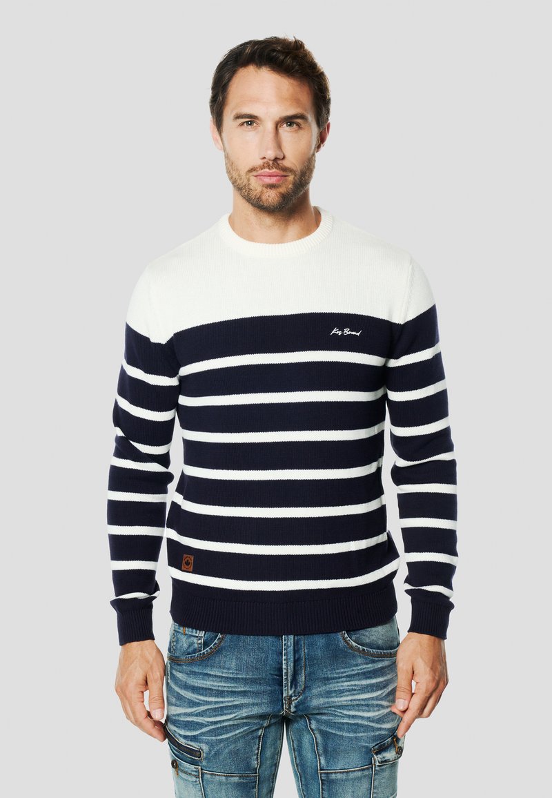 Marineblau-weiß gestreifter Strickpullover mit Rundhalsausschnitt, gerippten Bündchen und kleinem Logo auf der Brust. Kombiniert mit blauen Jeans.