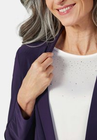 Lila Blazer mit glatter Textur, kombiniert mit einem weißen Oberteil, das mit verstreuten Strasssteinen verziert ist. Details am Kragen und am Ausschnitt sind sichtbar.
