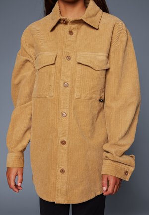 Beżowa sztruksowa koszula zapinana na guziki z dwoma przednimi kieszeniami z klapkami i długimi rękawami na osobie na jednolitym szarym tle.