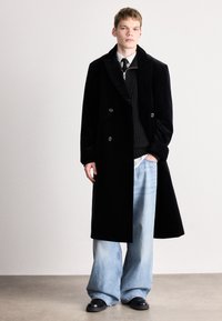 Μαύρο διπλό παλτό με γιακά σε στυλ σοφρού, φορεμένο πάνω από ένα μαύρο ριμπ πουλόβερ. Πλατιά μπλε τζιν παντελόνια και μαύρα παπούτσια ολοκληρώνουν την εικόνα.