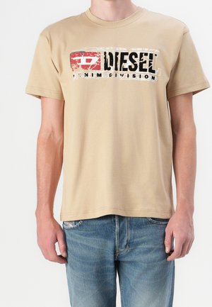 T-shirt Tan Diesel Denim Division porté par une personne en jean bleu, vue de face, montrant le logo et le texte sur la poitrine.