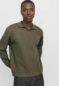 Polo de manga larga de color verde oliva, tejido, con un patrón texturizado y un cuello suave, combinado con vaqueros oscuros.