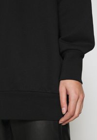 Sudadera negra con puños y dobladillo acanalados, que muestra una textura suave y un ajuste relajado. Una mano está situada cerca de la parte inferior de la prenda.
