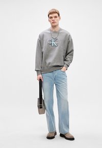 Šedá mikina s modrým logem, světle modré džíny rovného střihu, šedá obuv a malá šedá taška s černým popruhem.