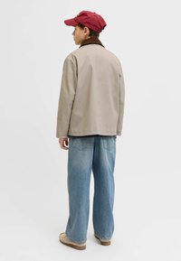 Jeune personne portant une veste beige, un jean bleu clair, des chaussures beiges et une casquette rouge, debout de profil devant un fond uni.