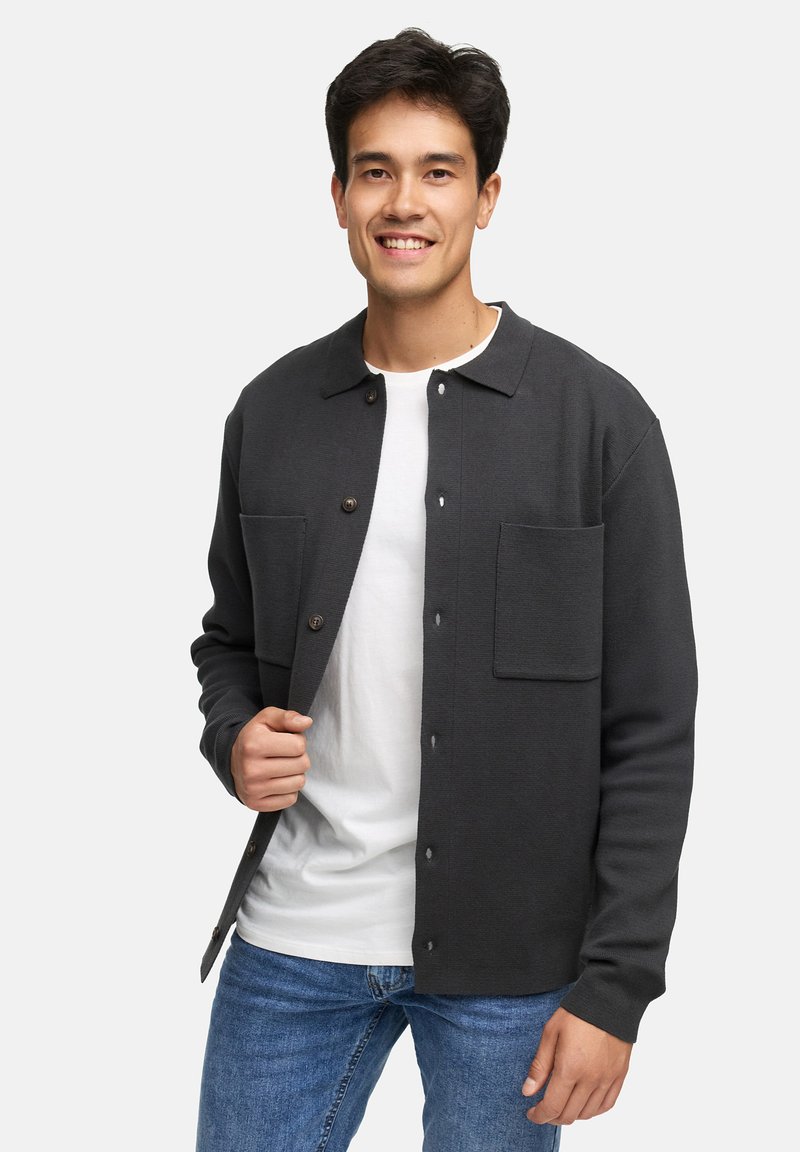 Chemise noire à boutonner en tissu doux avec deux poches poitrine, un col et une coupe décontractée, portée par-dessus un T-shirt blanc.